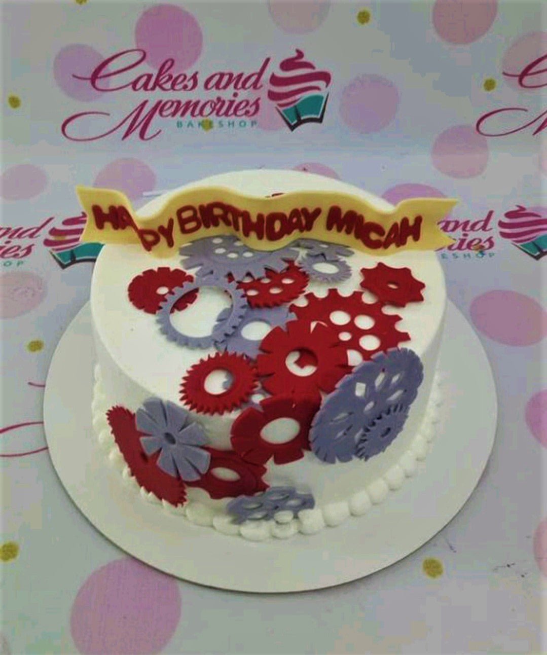 White round cake with red and purple gear decorations and a yellow banner message for Micah's birthday. White round cake with red and purple gear decorations and a yellow banner message for Micah's birthday.