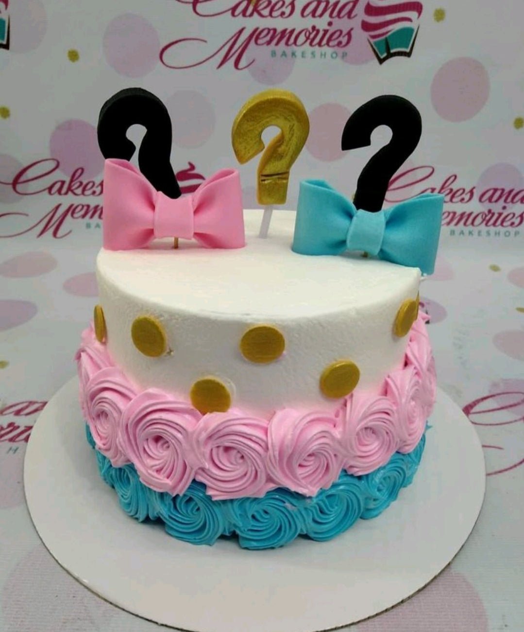 Gender reveal cake with white icing, pink and blue rosettes, gold dots, and question mark toppers with bows. Gender reveal cake with white icing, pink and blue rosettes, gold dots, and question mark toppers with bows.