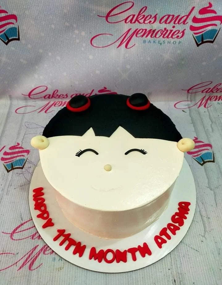 Girl Face White Cake A white 1-tier round cake themed as a girl's face, featuring black fondant hair, red hair ties, facial features, and a message on the base board.