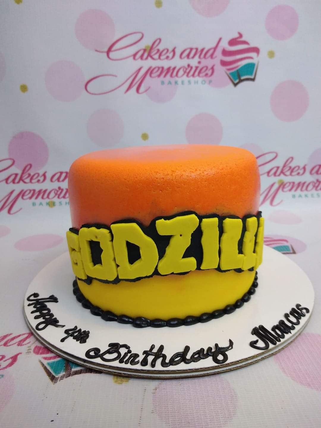 A single-tier Godzilla themed birthday cake with orange and yellow gradient icing and a large yellow Godzilla logo on the side. A single-tier Godzilla themed birthday cake with orange and yellow gradient icing and a large yellow Godzilla logo on the side.