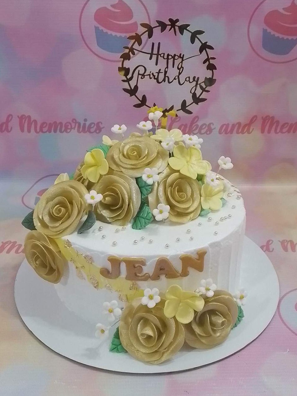 White floral birthday cake for Jean featuring gold roses, yellow blossoms, and a Happy Birthday wreath topper. White floral birthday cake for Jean featuring gold roses, yellow blossoms, and a Happy Birthday wreath topper.