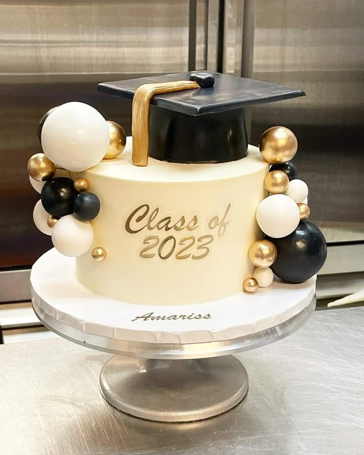 White graduation cake with a black fondant graduation cap, gold tassel, and a cluster of black, white, and gold decorative spheres. White graduation cake with a black fondant graduation cap, gold tassel, and a cluster of black, white, and gold decorative spheres.