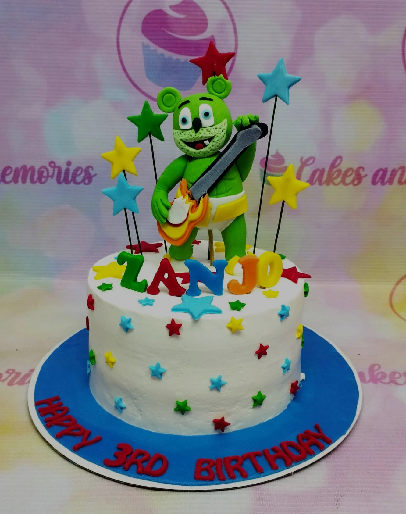 White soft-icing Gummy Bear themed cake with a large green fondant bear playing guitar and colorful fondant stars. White soft-icing Gummy Bear themed cake with a large green fondant bear playing guitar and colorful fondant stars.