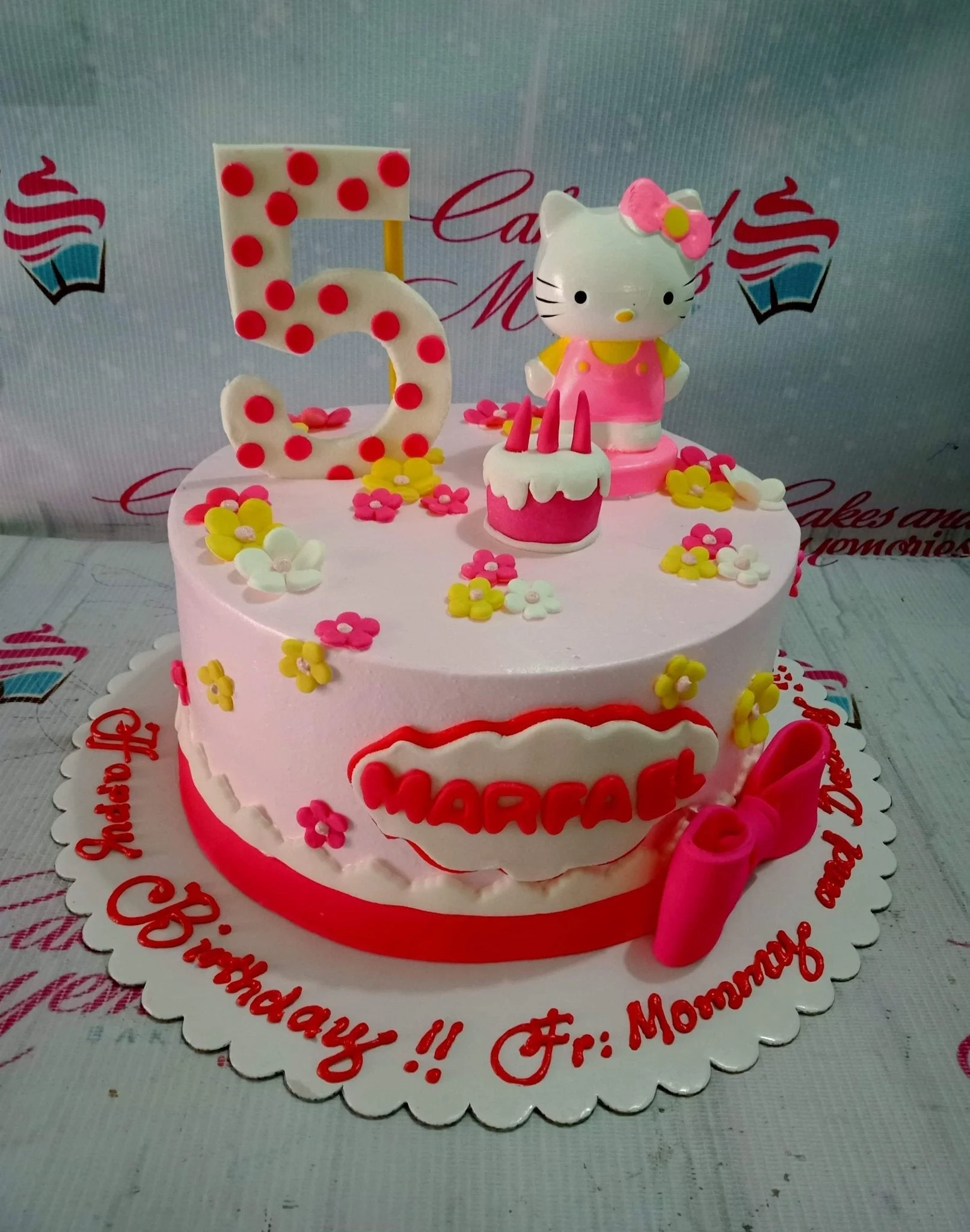 Pink Hello Kitty birthday cake with a large polka dot number 5, fondant flowers, and a pink bow on a white scalloped board.