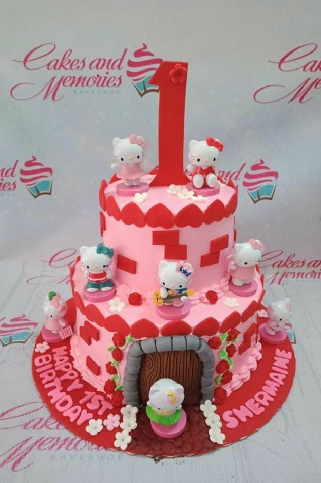 Two-tier pink Hello Kitty themed birthday cake with multiple character figurines, red hearts, and a large red number one topper. Two-tier pink Hello Kitty themed birthday cake with multiple character figurines, red hearts, and a large red number one topper.