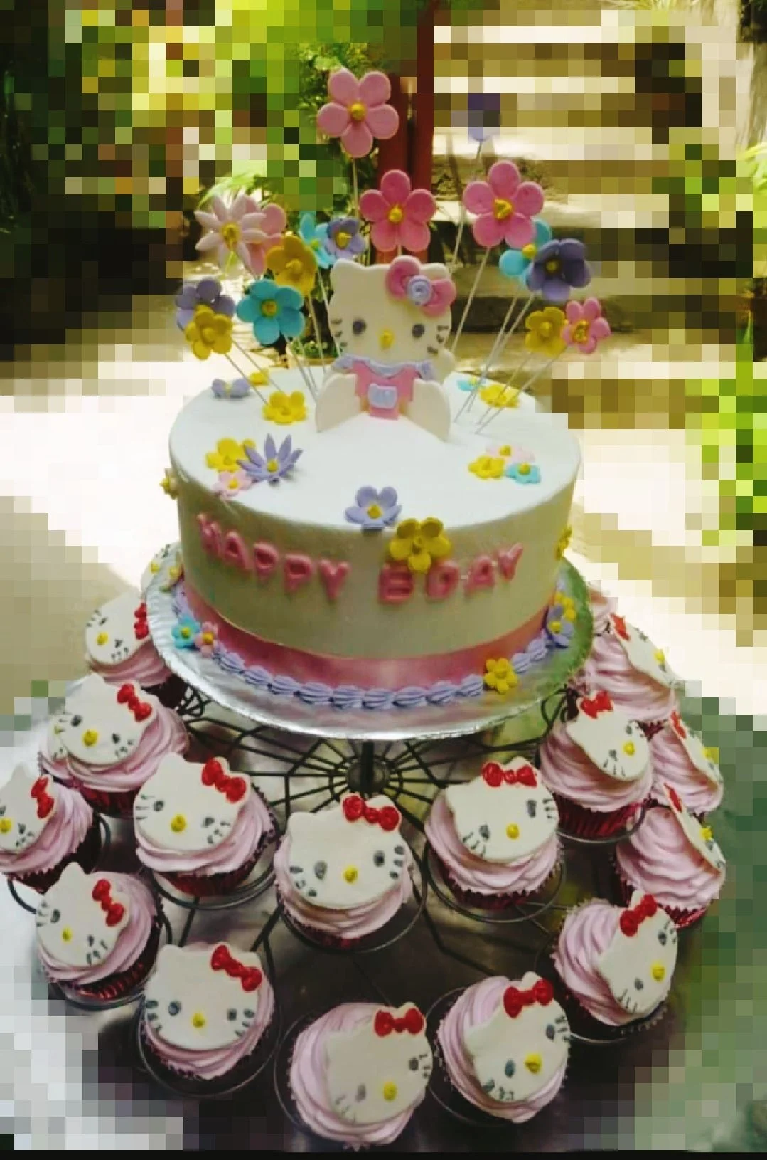 Hello Kitty themed birthday cake with pink and purple icing rosettes, fondant flowers, and a Hello Kitty topper surrounded by cupcakes.