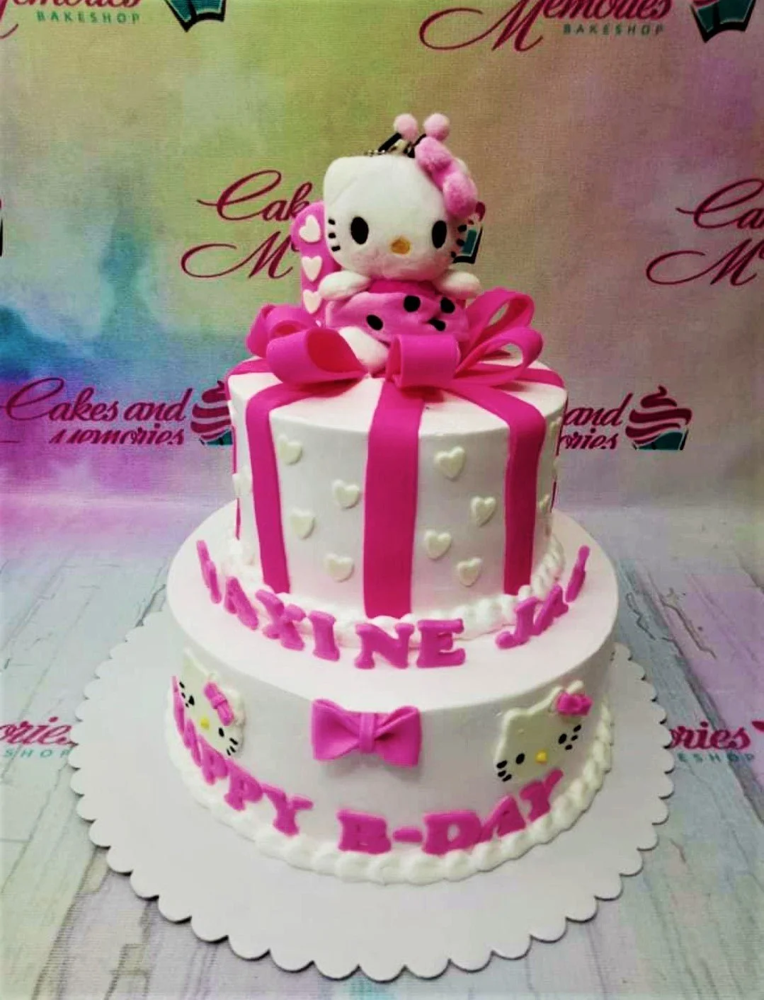 Two-tier white Hello Kitty themed cake with pink ribbons, hearts, and a plush Hello Kitty topper.