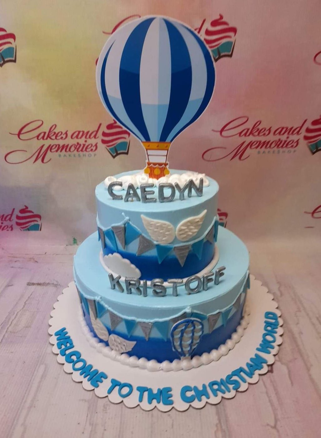 Two-tier light blue hot air balloon baptism cake with silver gumpaste letters and blue bunting flags. Two-tier light blue hot air balloon baptism cake with silver gumpaste letters and blue bunting flags.