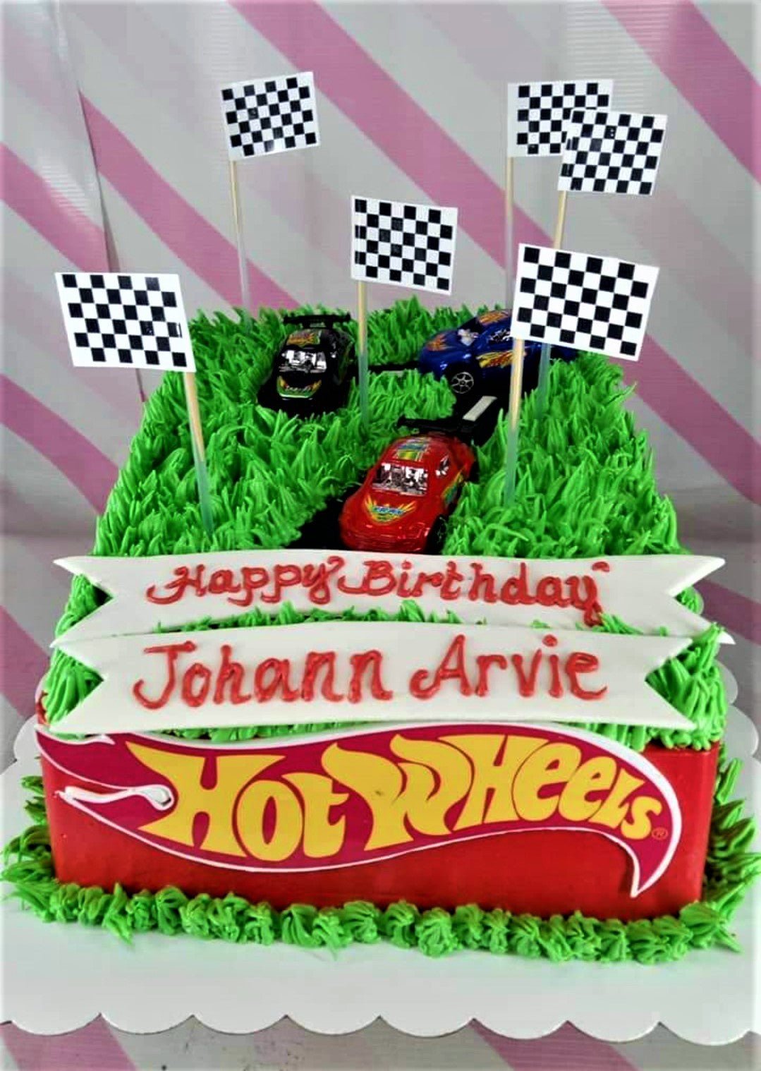 Square Hot Wheels themed birthday cake with green grass icing, toy cars, checkered flags, and a personalized name banner. Square Hot Wheels themed birthday cake with green grass icing, toy cars, checkered flags, and a personalized name banner.