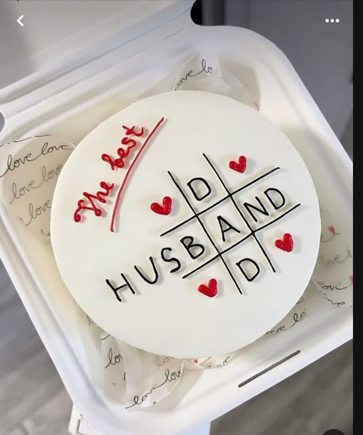White bento cake with a tic-tac-toe design featuring red hearts and the message 'The best HUSBAND' in black and red icing. White bento cake with a tic-tac-toe design featuring red hearts and the message 'The best HUSBAND' in black and red icing.