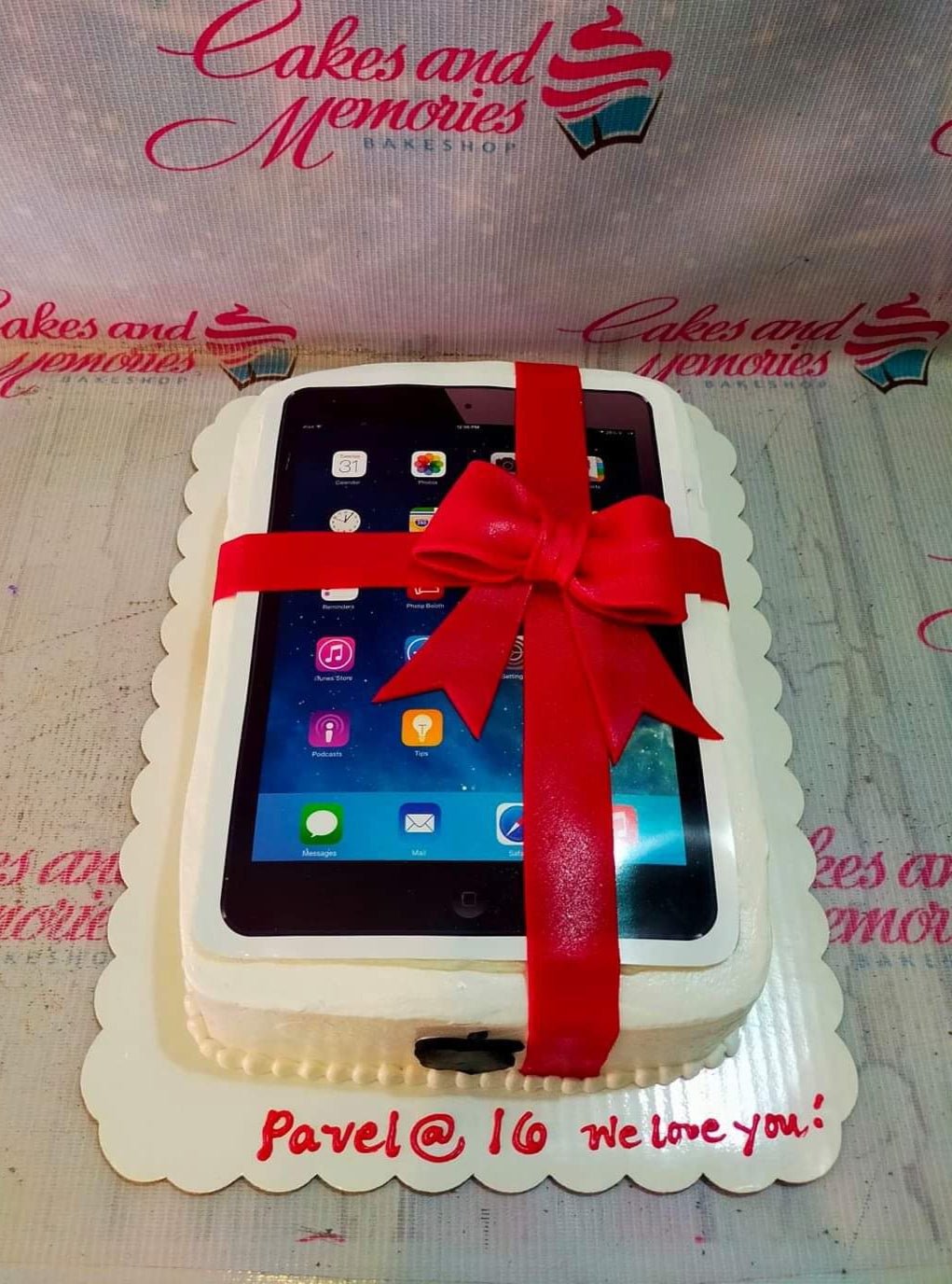White rectangular cake designed to look like an iPad with a large red fondant bow and a printed screen showing app icons. White rectangular cake designed to look like an iPad with a large red fondant bow and a printed screen showing app icons.