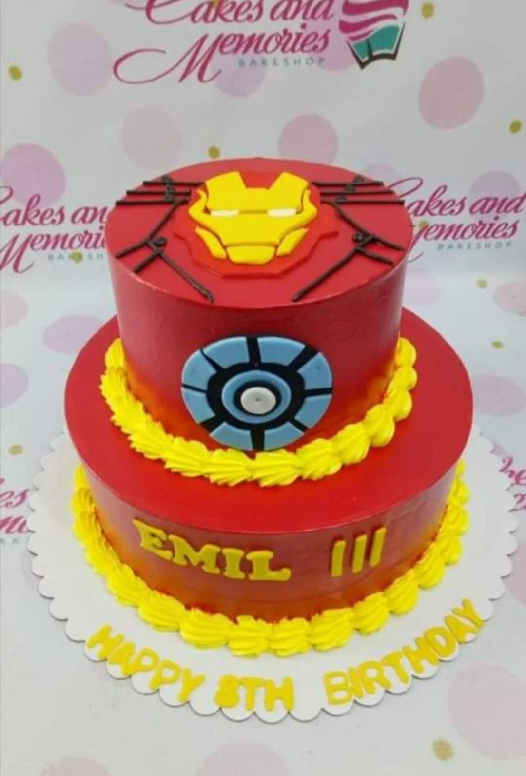 Iron Man-Inspired 2-Tier Birthday Cake Two-tier red Iron Man themed birthday cake with yellow icing borders and fondant superhero emblems.