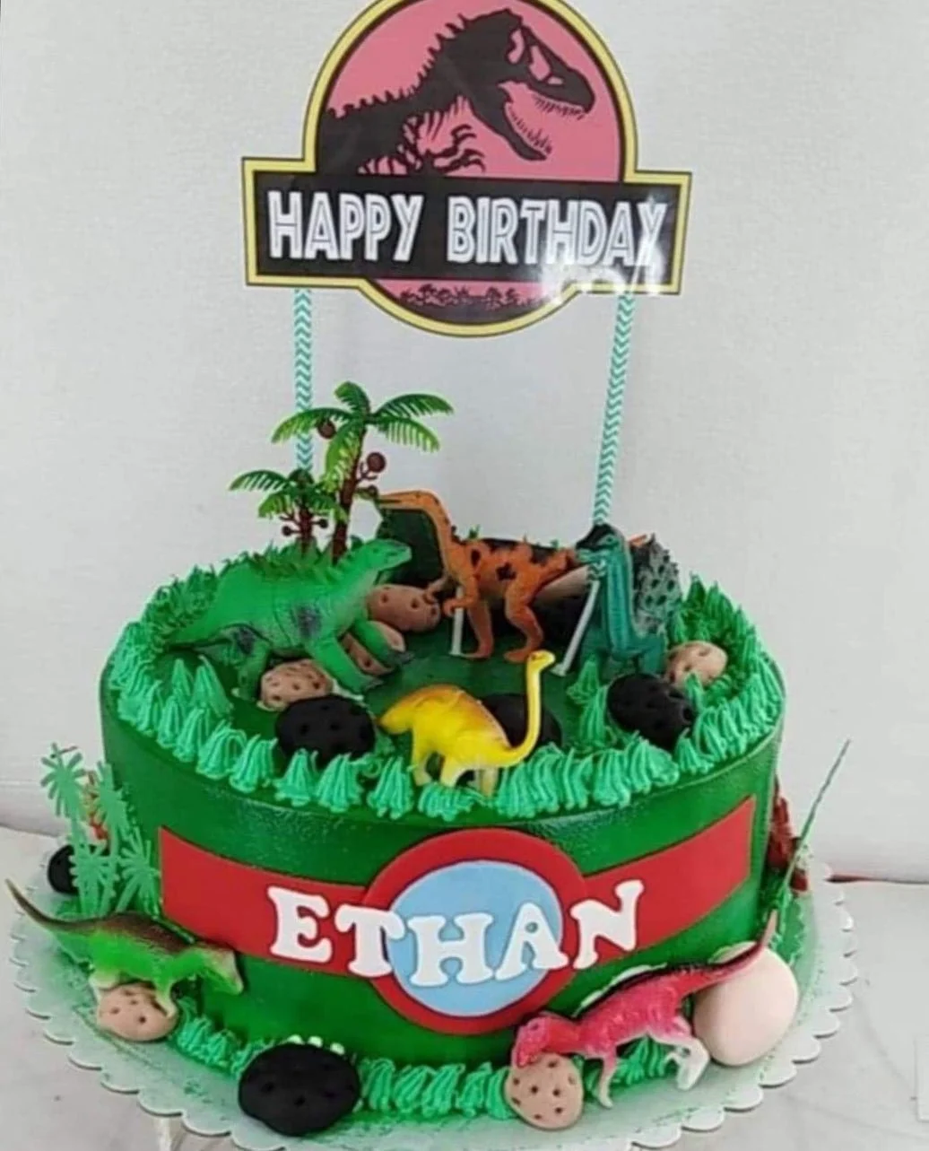 Green Jurassic Park themed dinosaur birthday cake with plastic dinosaur toys, palm trees, and a custom Ethan name banner.