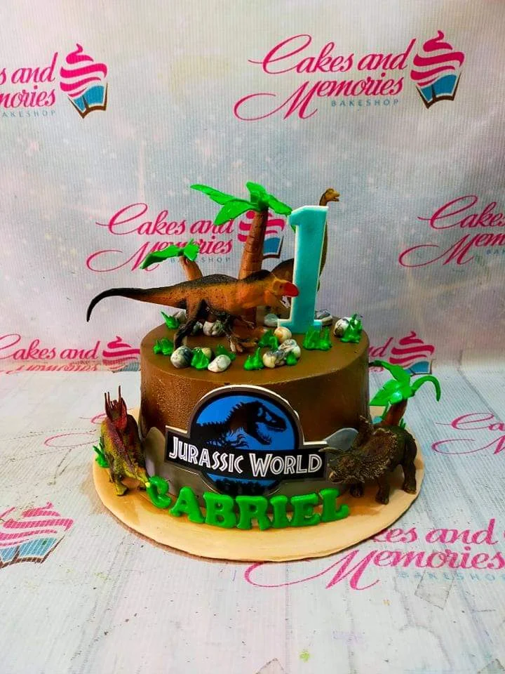 Brown Jurassic World dinosaur cake with 3D dinosaur toys, palm trees, and a blue number one candle for Gabriel's first birthday.