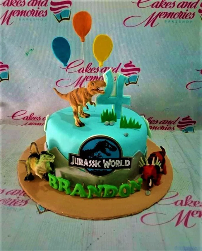 Blue Jurassic World dinosaur cake with a T-Rex topper, balloons, and green gumpaste name Brandon on a brown base board.