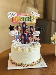 White soft-icing cake with colorful sprinkles at the base and multiple K-pop group printout toppers including a large Gabby name banner. White soft-icing cake with colorful sprinkles at the base and multiple K-pop group printout toppers including a large Gabby name banner.