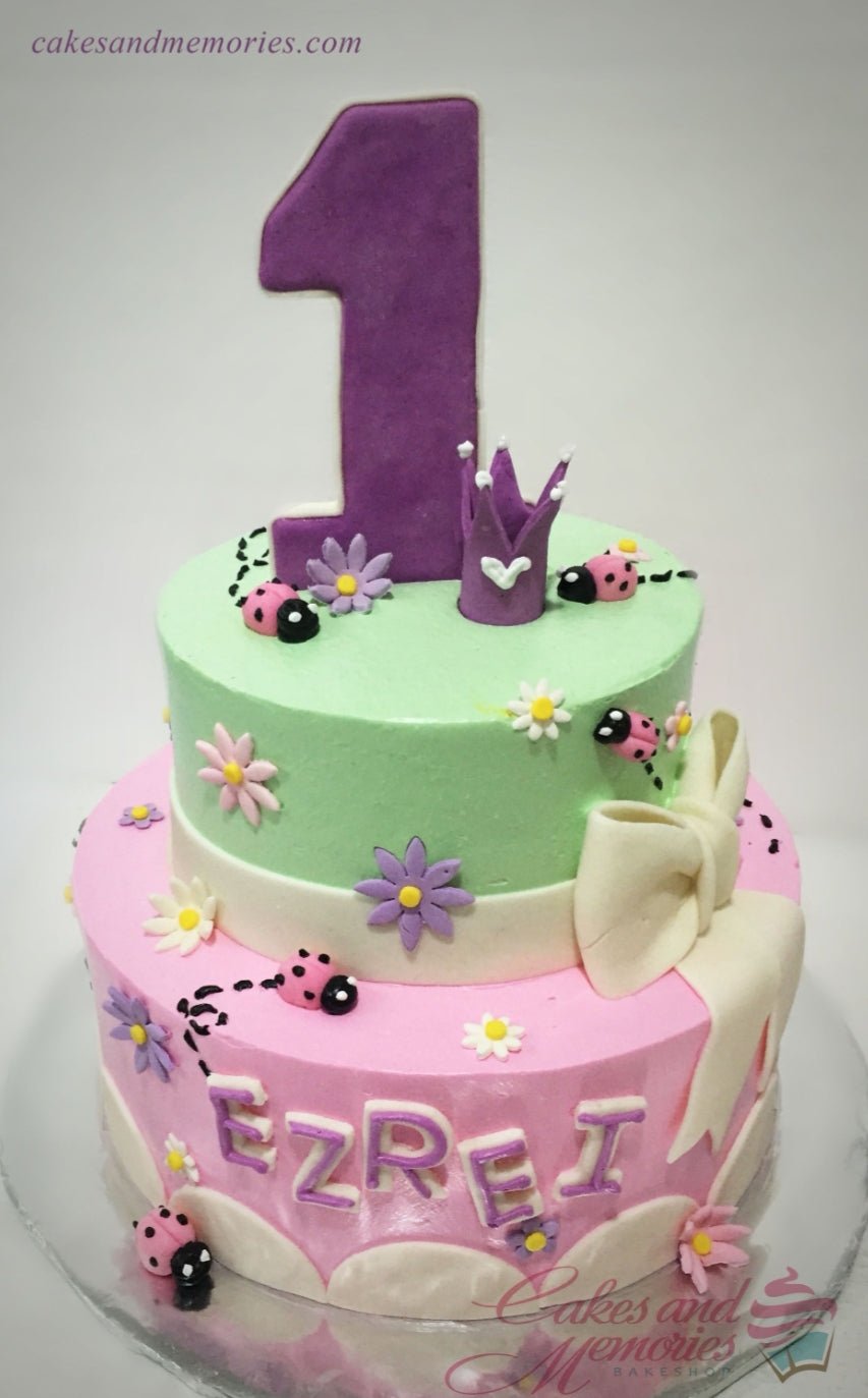 Two-tier pink and green garden-themed cake with ladybugs, flowers, a large purple number one, and a crown topper. Two-tier pink and green garden-themed cake with ladybugs, flowers, a large purple number one, and a crown topper.