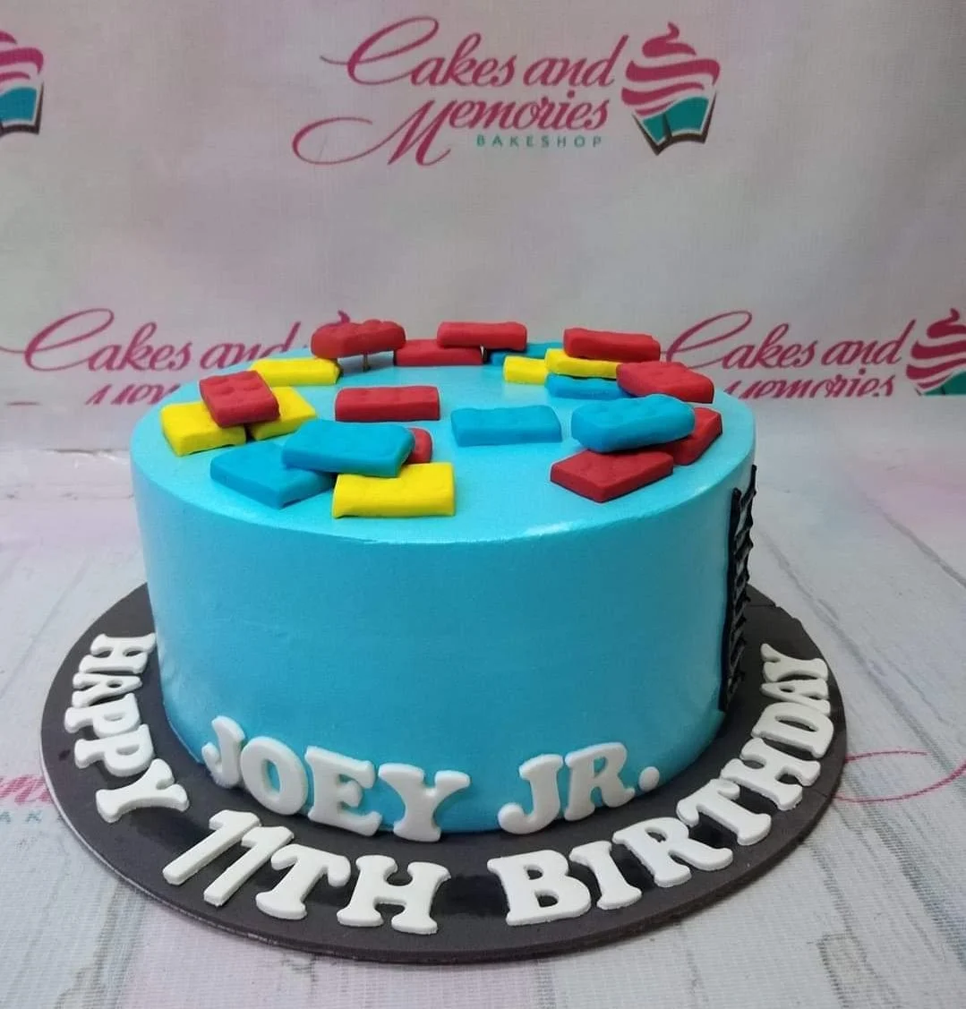 Lego-Inspired Sky Blue Birthday Cake A light blue and colorful 1 Tier cake themed around Lego, featuring fondant building blocks and white gumpaste letters on a black base board.