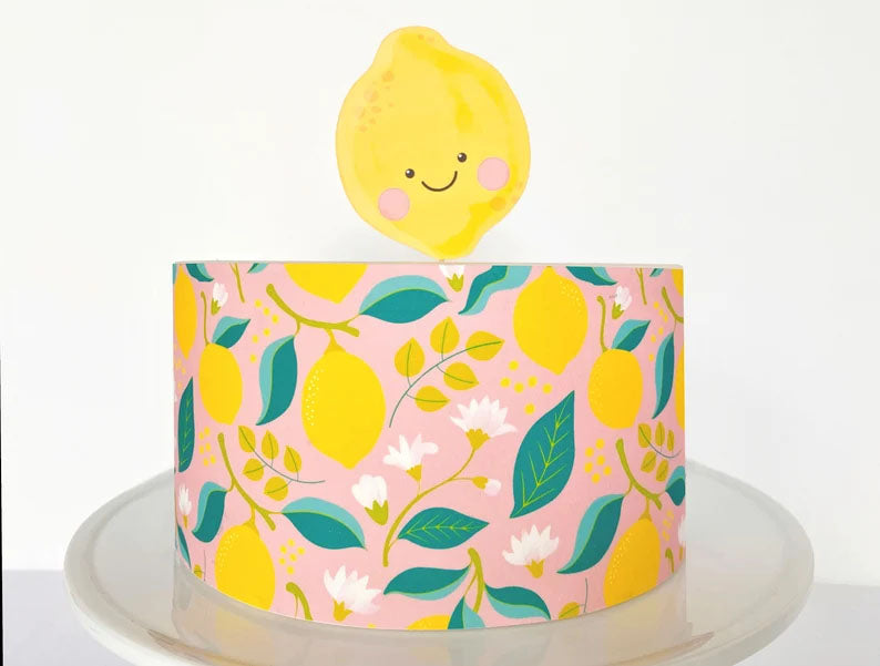 Single tier pink cake with a lemon pattern wrap and a smiling lemon character topper. Single tier pink cake with a lemon pattern wrap and a smiling lemon character topper.