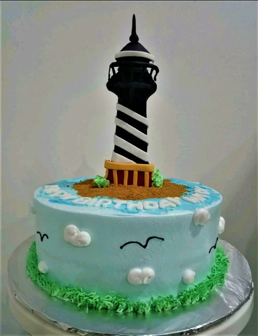 Lighthouse Sky Blue Birthday Cake A light blue 1 Tier lighthouse themed cake, featuring a black and white lighthouse on an island, with piped clouds, grass, and birds.