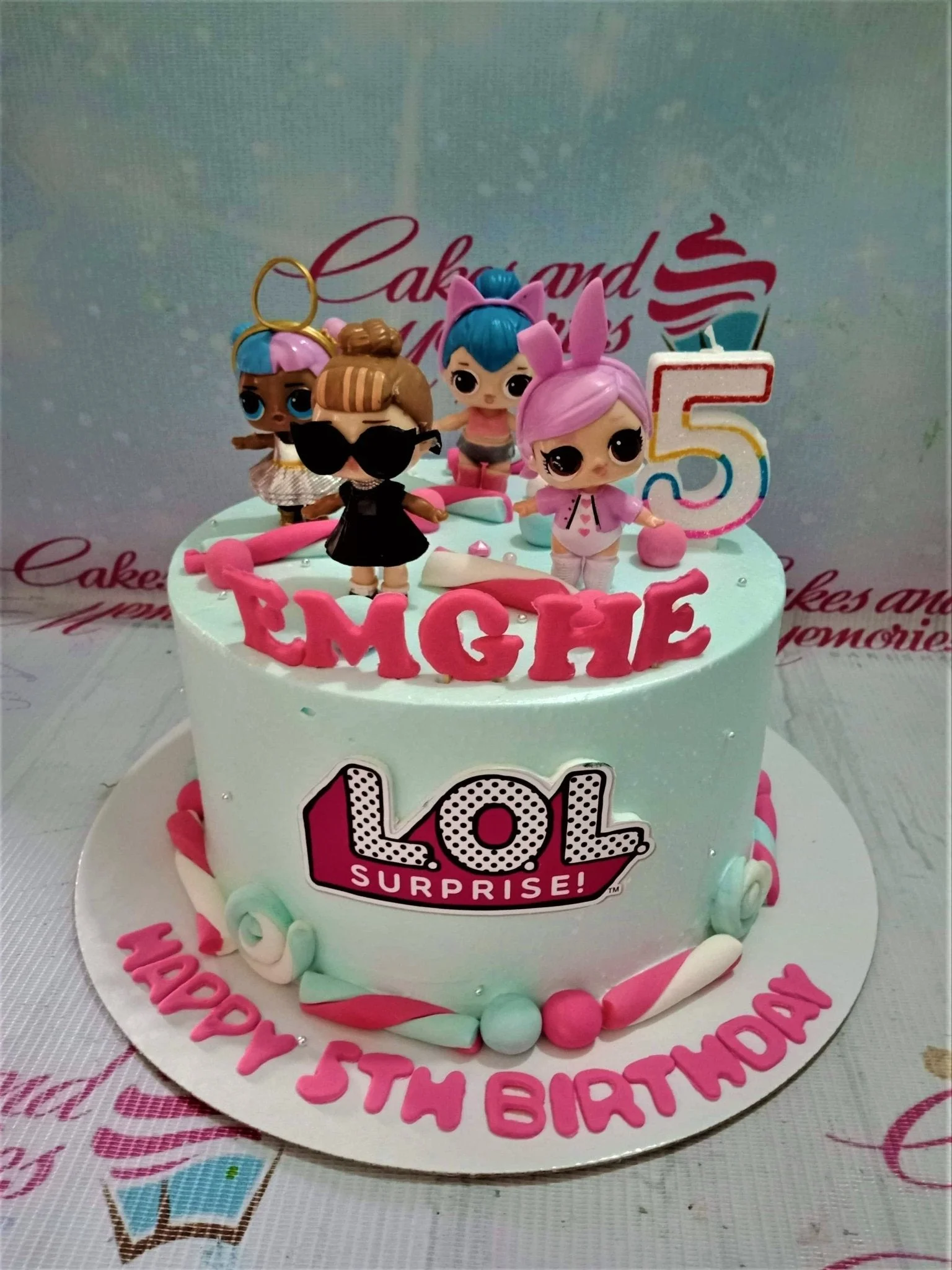 Light blue LOL Surprise themed birthday cake with 3D character toys, pink gumpaste letters, and marshmallow-style icing decorations.