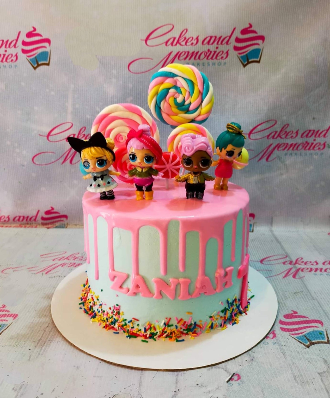 Light blue 1-tier LOL Surprise themed birthday cake with pink drip, colorful lollipops, and character toys. Light blue 1-tier LOL Surprise themed birthday cake with pink drip, colorful lollipops, and character toys.