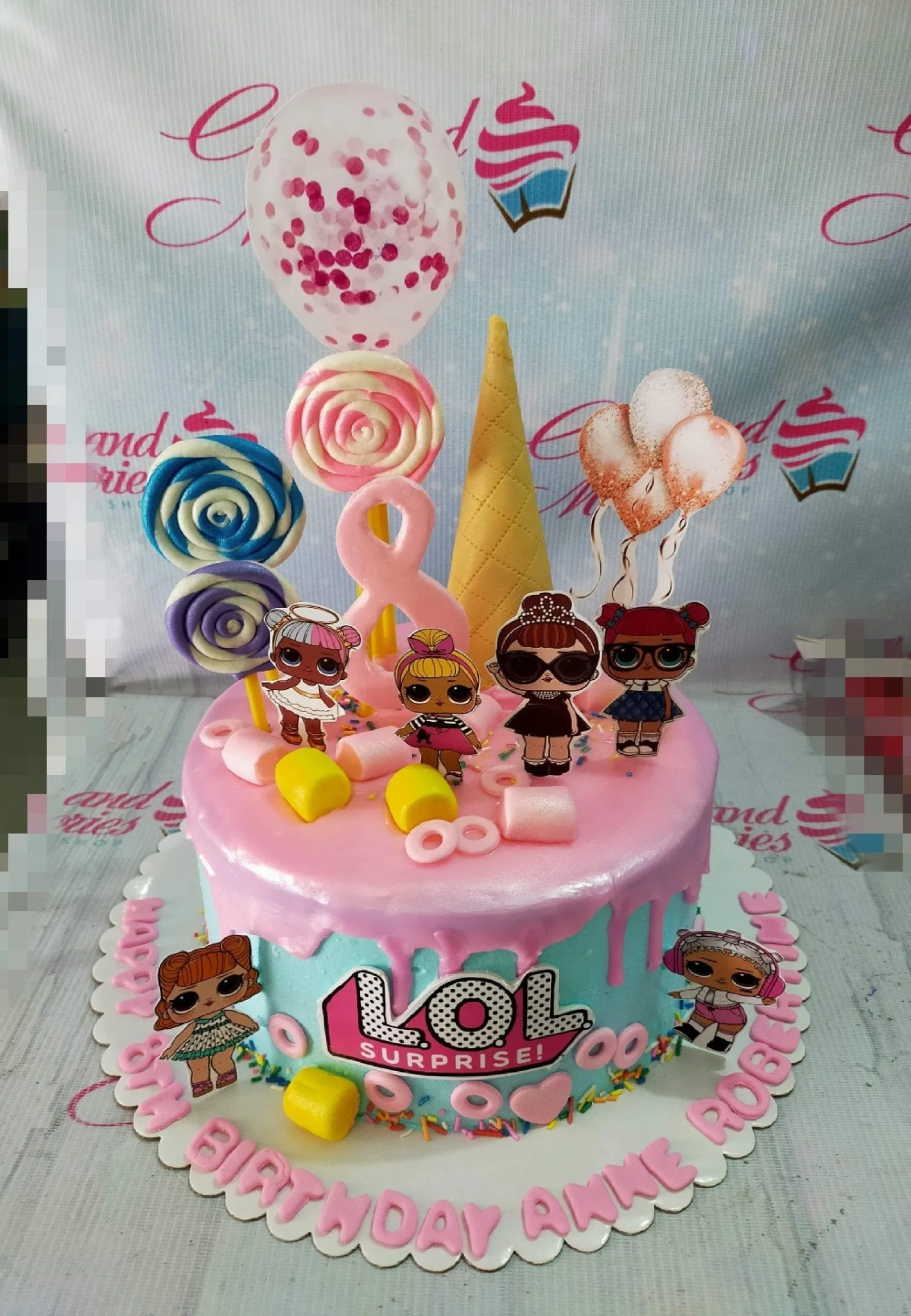LOL Surprise themed birthday cake with pink drip icing, character printouts, lollipops, and marshmallows on a light blue base.