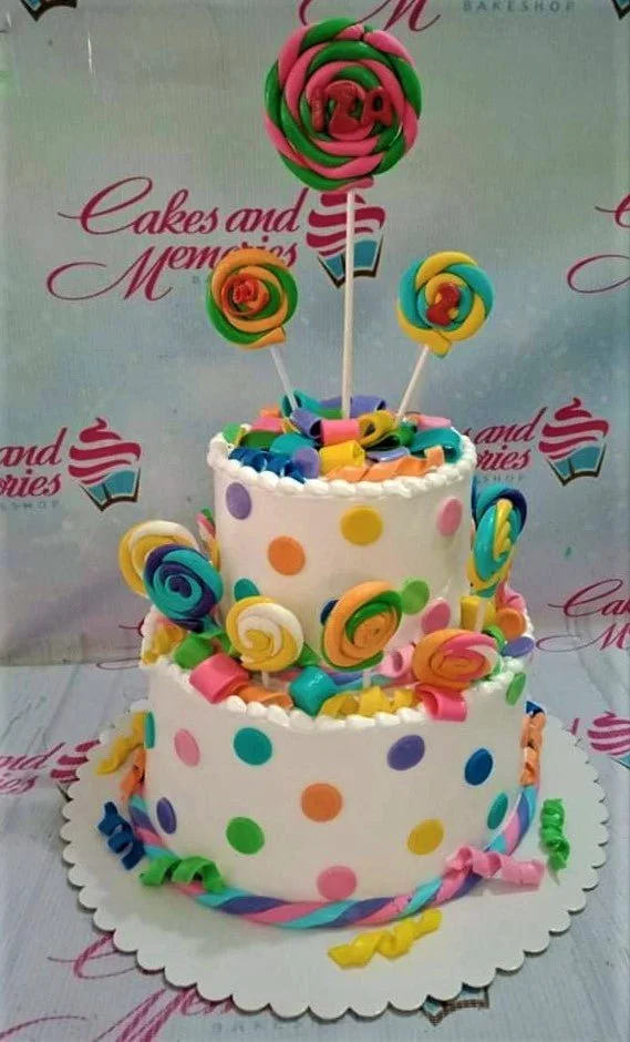 Lollipop White 2-Tier Birthday Cake Colorful 2-tier lollipop themed birthday cake with white icing, multi-colored polka dots, and fondant lollipop toppers.