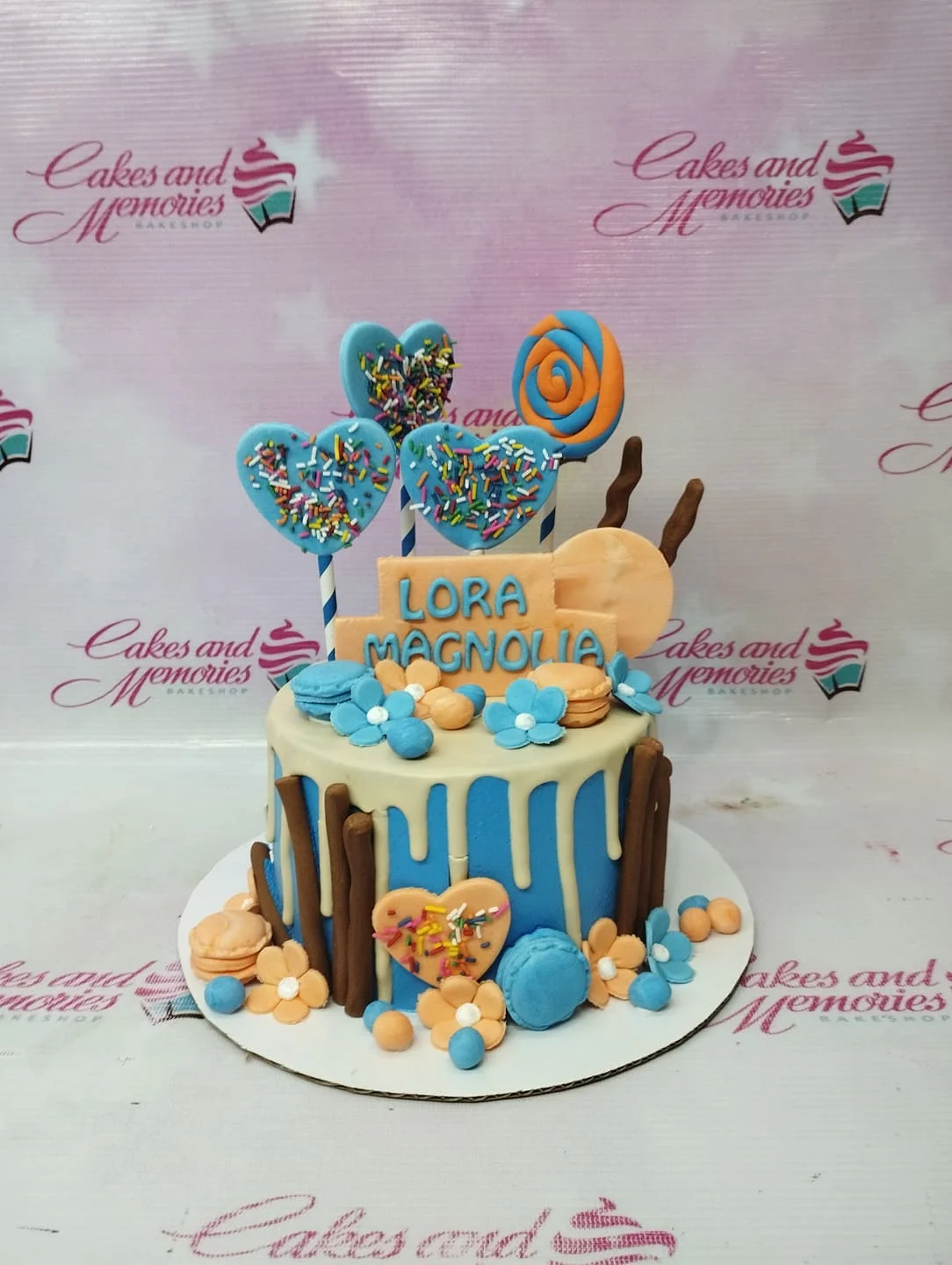 Lora Magnolia Blue Drip Birthday Cake A blue and orange 1-tier cake themed for Lora Magnolia, featuring heart-shaped toppers, a swirl lollipop, chocolate sticks, and macarons.
