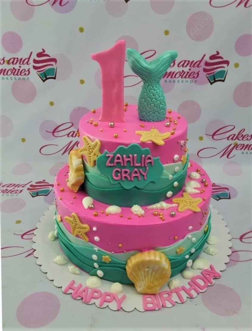 Pink and teal 2-tier mermaid themed birthday cake with a large pink number one and a teal mermaid tail topper. Pink and teal 2-tier mermaid themed birthday cake with a large pink number one and a teal mermaid tail topper.