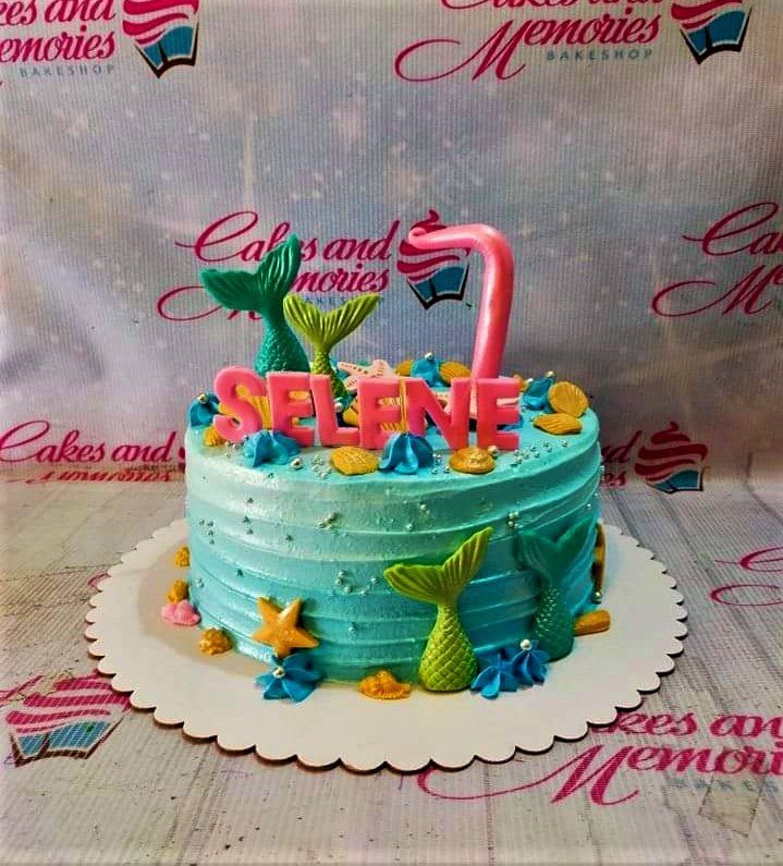 Light blue mermaid themed birthday cake with pink name topper, mermaid tails, and gold seashells. Light blue mermaid themed birthday cake with pink name topper, mermaid tails, and gold seashells.