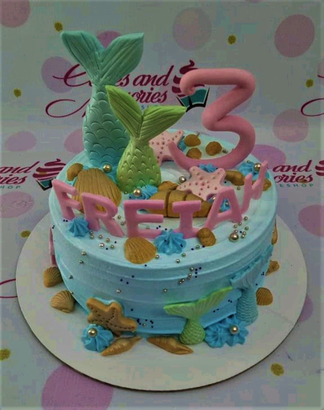 Light blue mermaid themed birthday cake with pink fondant letters, mermaid tails, and gold seashells. Light blue mermaid themed birthday cake with pink fondant letters, mermaid tails, and gold seashells.