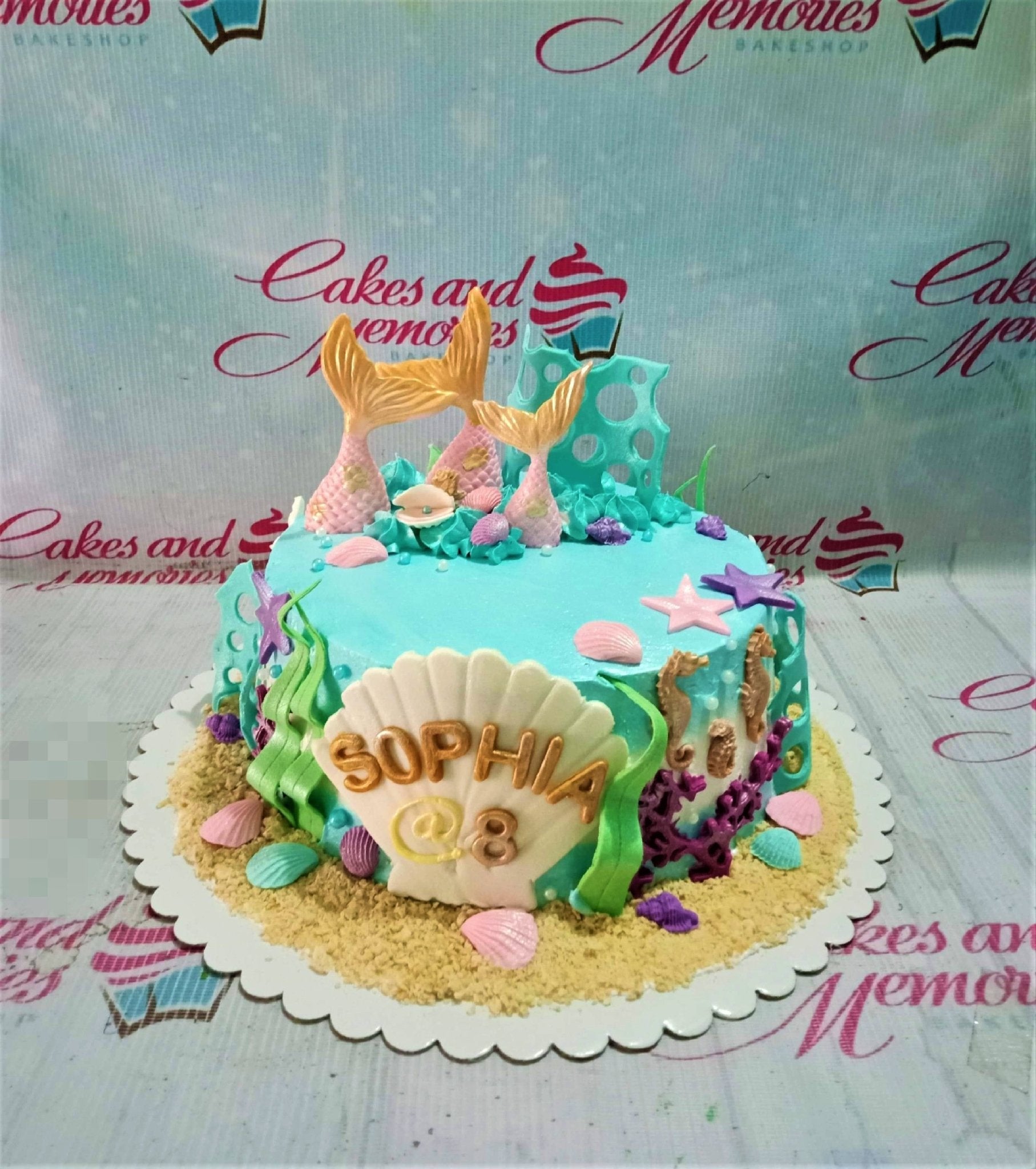Light blue mermaid themed birthday cake with gold mermaid tails, seashells, seahorses, and a large white shell with the name Sophia. Light blue mermaid themed birthday cake with gold mermaid tails, seashells, seahorses, and a large white shell with the name Sophia.