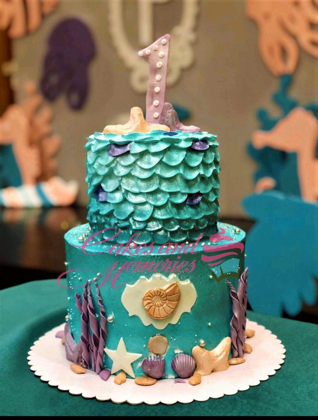Two-tier mermaid themed cake in teal and purple with edible sea shells, seaweed, and a number one topper. Two-tier mermaid themed cake in teal and purple with edible sea shells, seaweed, and a number one topper.