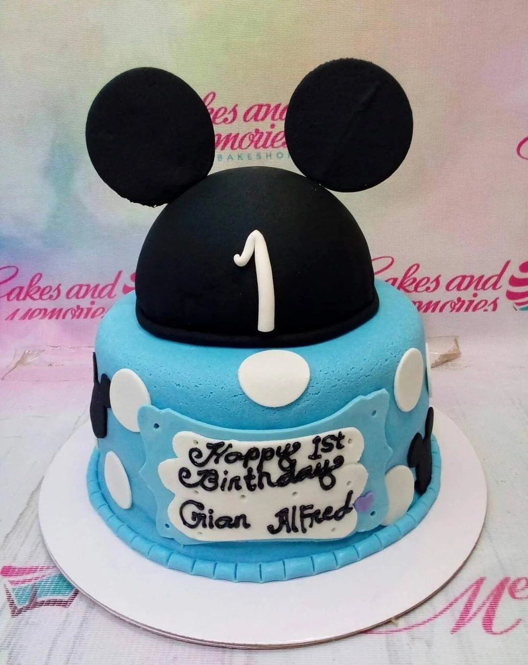 Blue Mickey Mouse themed 1st birthday cake with a black fondant hat topper and white polka dots.