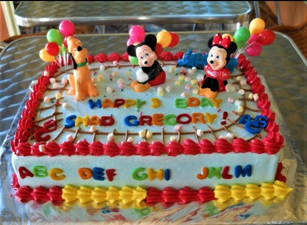 A light blue rectangular Mickey Mouse themed cake, featuring Mickey, Minnie, Pluto figurines, colorful balloons, a blue toy train, and icing text. A light blue rectangular Mickey Mouse themed cake, featuring Mickey, Minnie, Pluto figurines, colorful balloons, a blue toy train, and icing text.