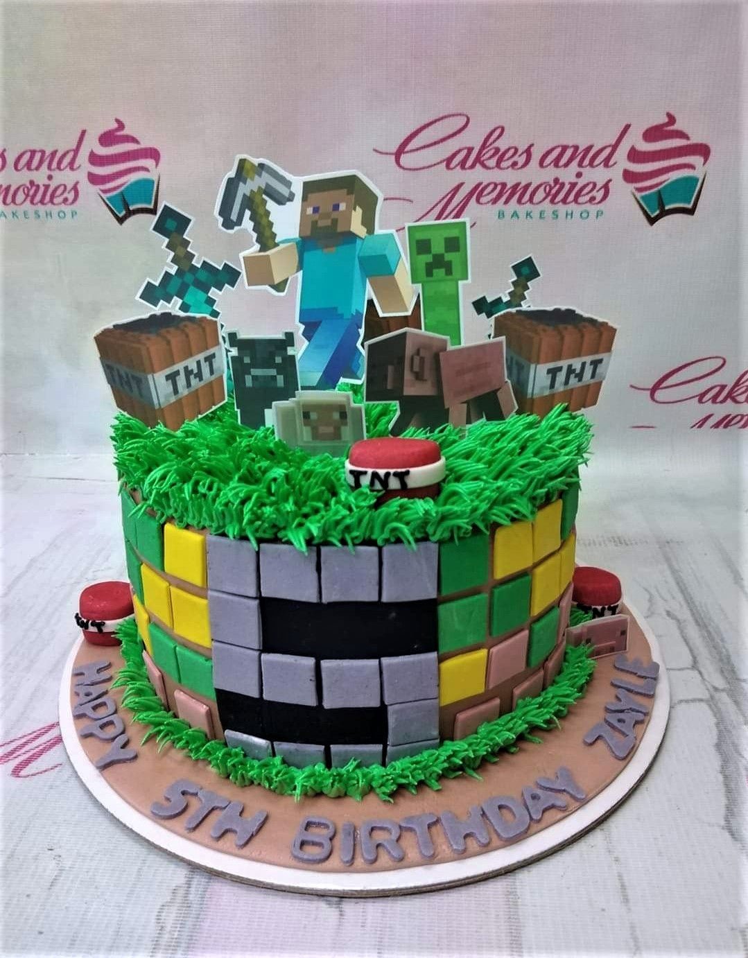 Minecraft themed birthday cake with Steve, Creeper, and TNT toppers on green grass icing with a block-patterned side design.