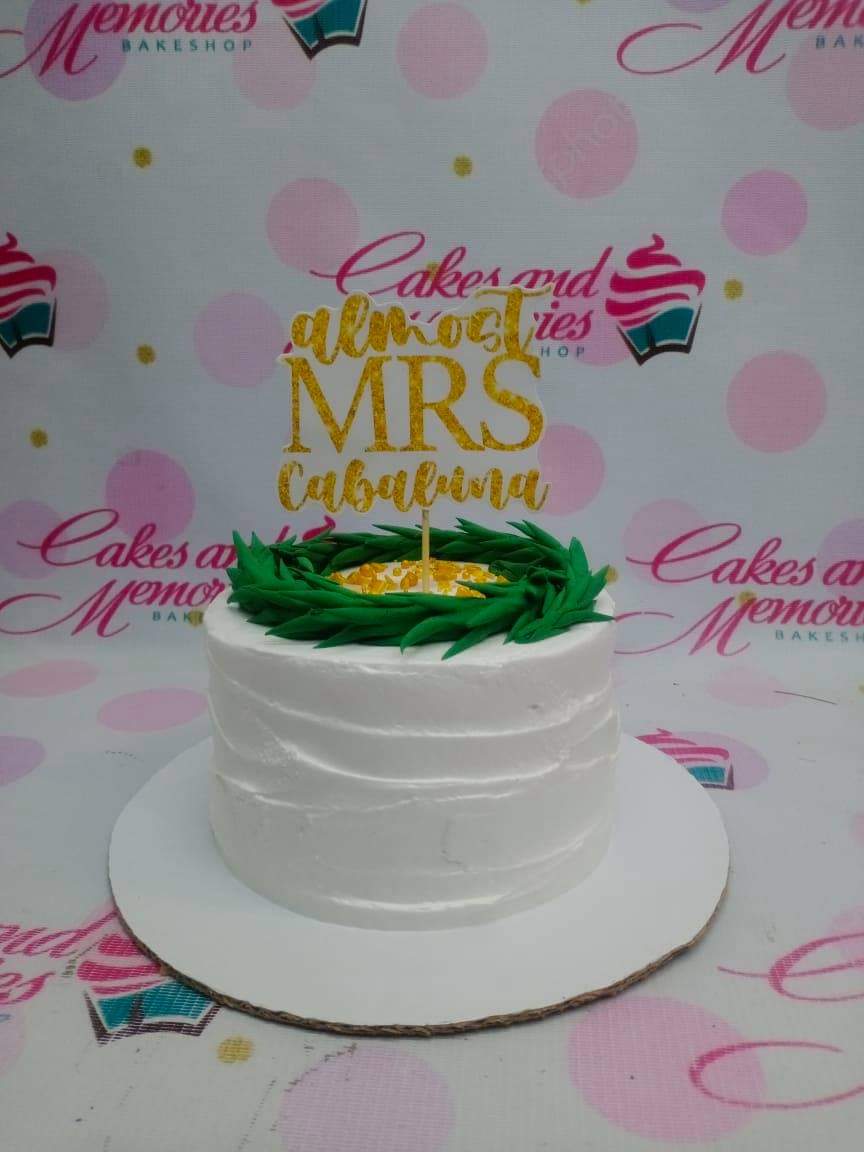 White minimalist cake with textured icing, a green leaf wreath on top, and a gold glittery Mrs Cabaluna topper.