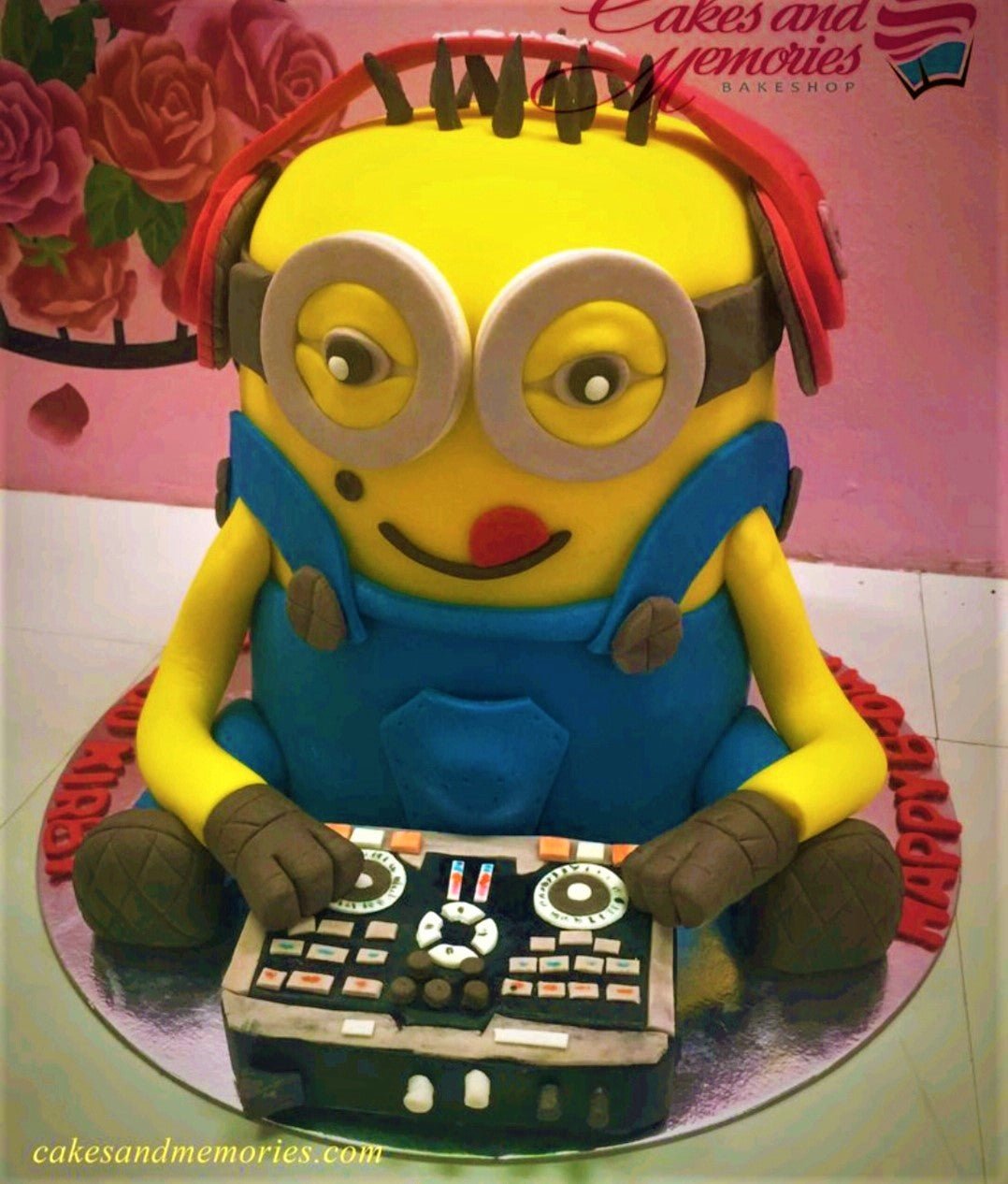 Custom 3D Minion DJ cake with headphones and a detailed mixing console on a silver base board. Custom 3D Minion DJ cake with headphones and a detailed mixing console on a silver base board.