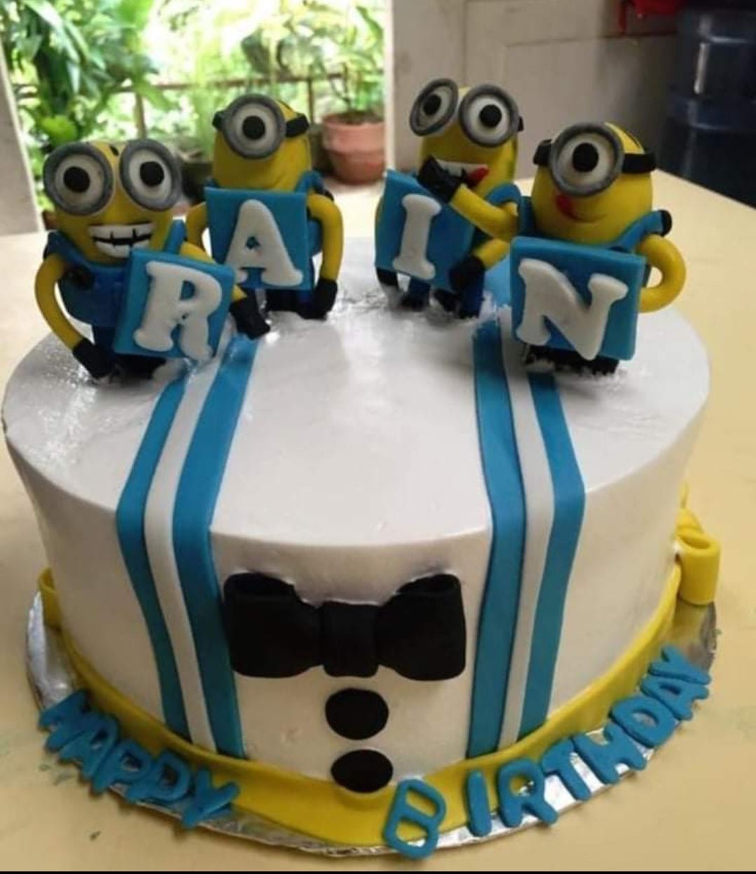 White 1-tier Minion themed birthday cake with blue stripes, a black fondant bow tie, and four 3D Minion figures holding letters. White 1-tier Minion themed birthday cake with blue stripes, a black fondant bow tie, and four 3D Minion figures holding letters.