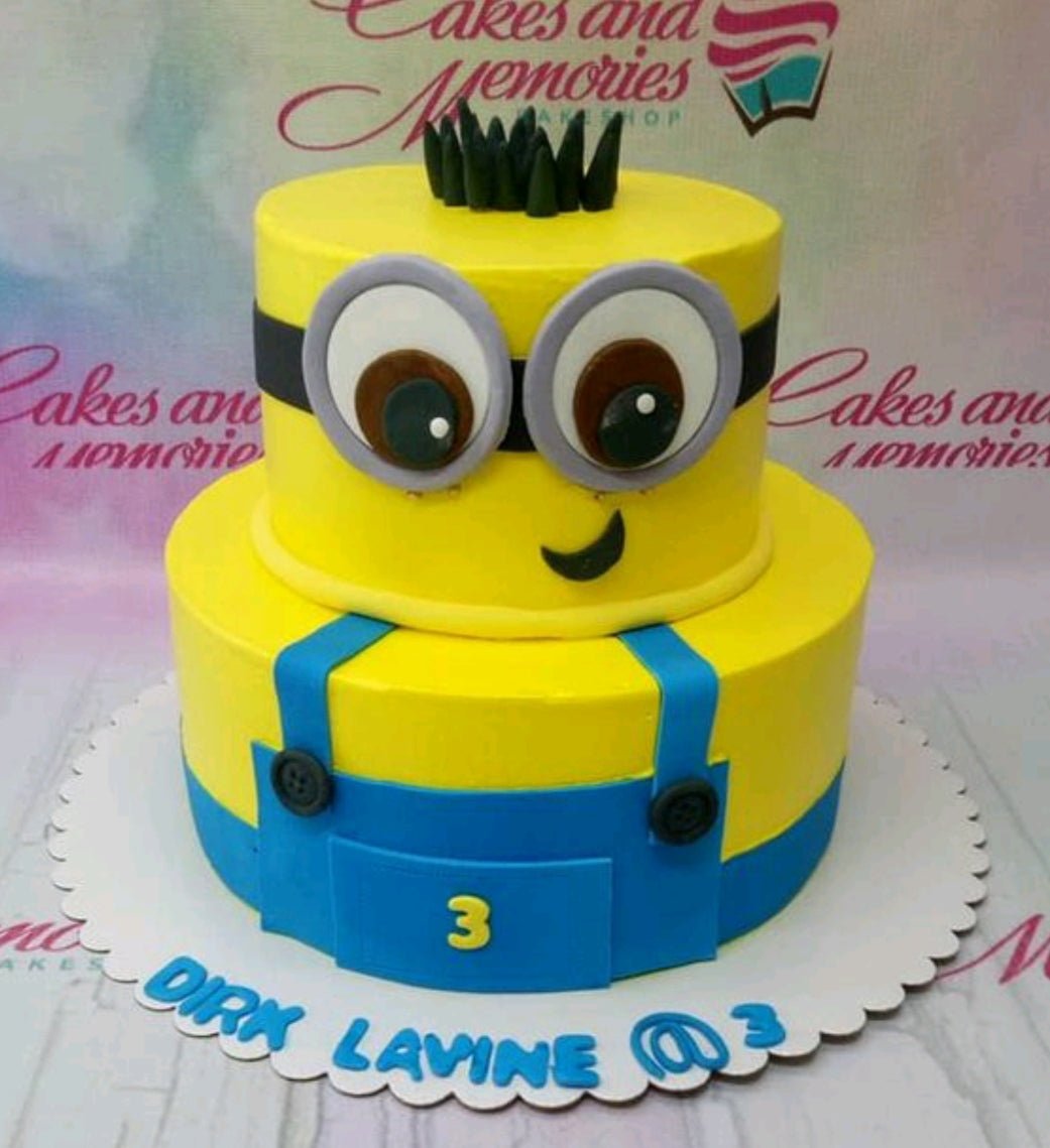 Two-tier yellow Minion themed cake with blue overalls, gumpaste eyes, and black hair accents. Two-tier yellow Minion themed cake with blue overalls, gumpaste eyes, and black hair accents.