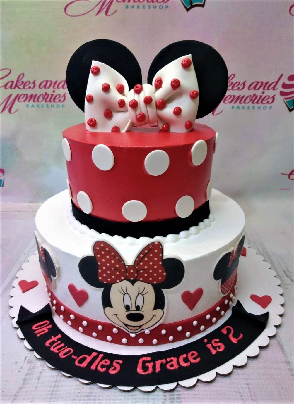 Two-tier Minnie Mouse themed birthday cake in red, white, and black with a large fondant bow and character printouts. Two-tier Minnie Mouse themed birthday cake in red, white, and black with a large fondant bow and character printouts.