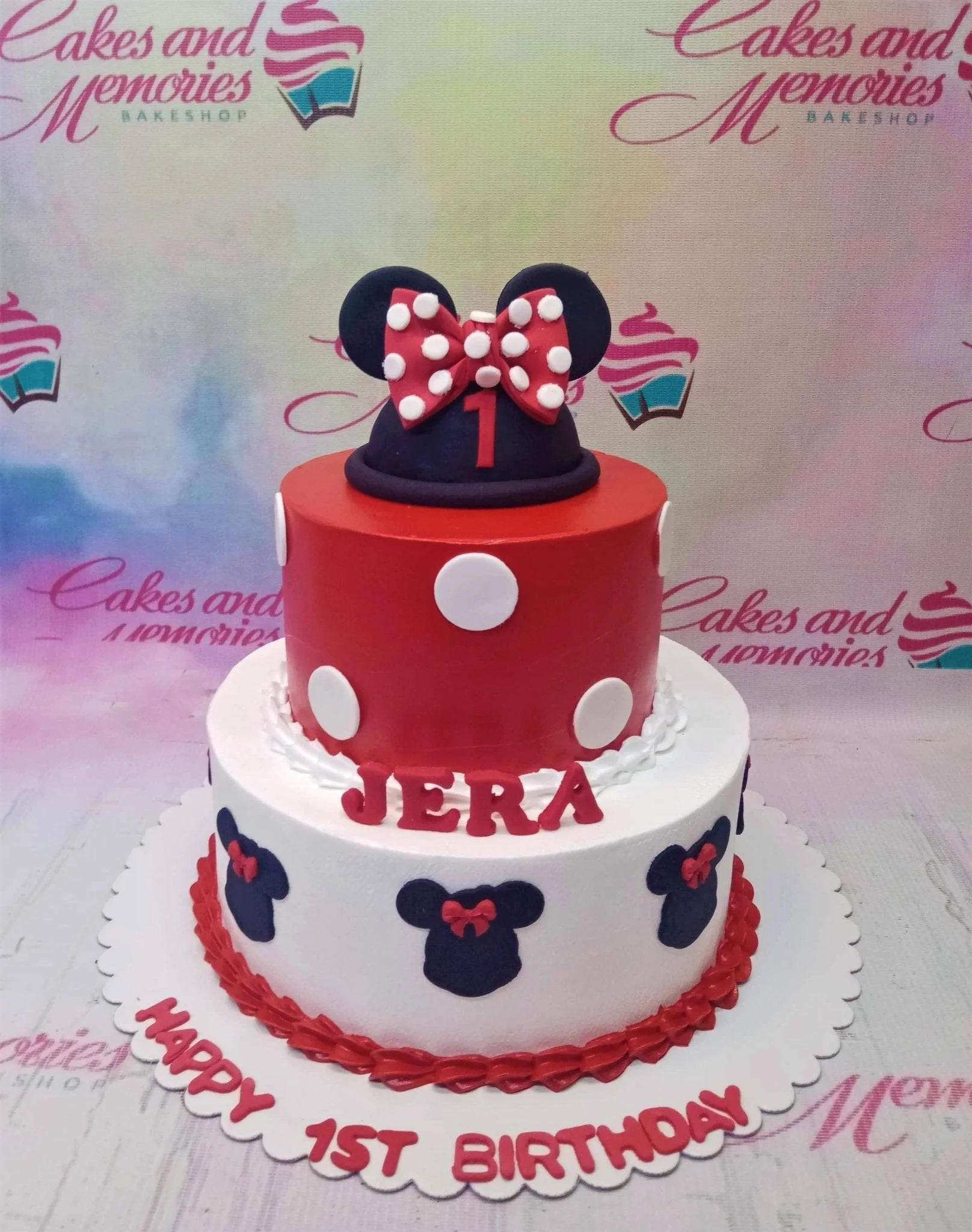 A red, white, and black two-tier Minnie Mouse cake, with mouse ears topper, polka dot bow, and mouse silhouettes.