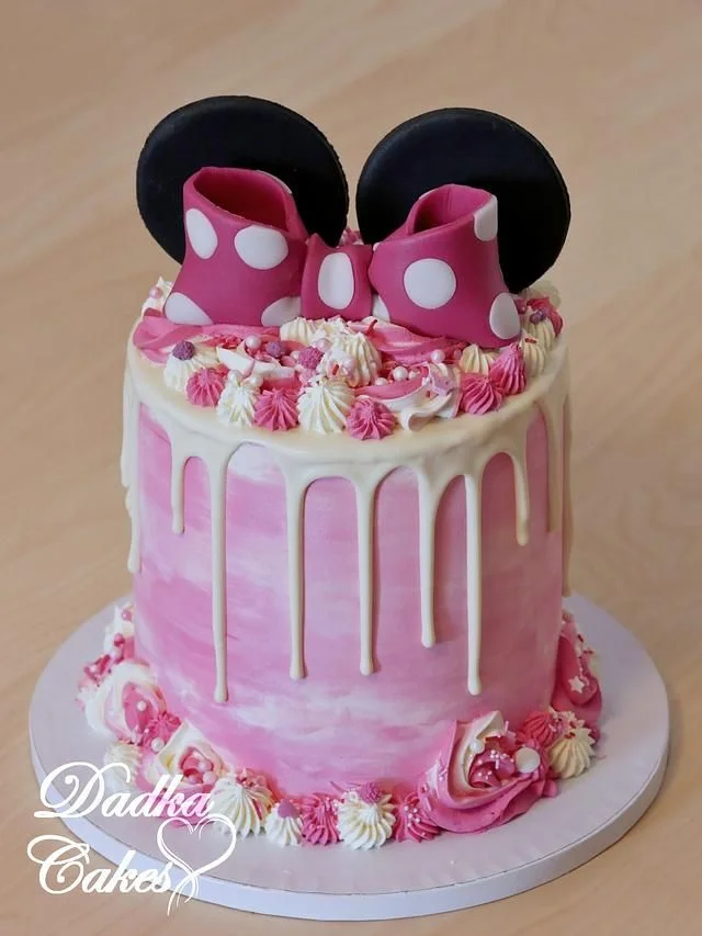 Pink Minnie Mouse themed cake with white chocolate drip, pink and white icing rosettes, and large fondant Minnie ears and bow topper.