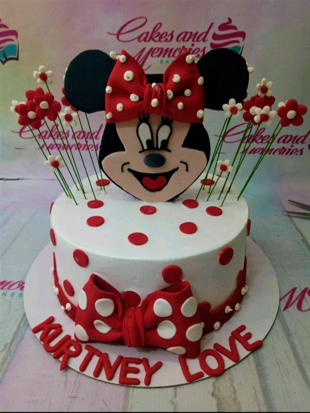 White Minnie Mouse themed cake with red polka dots, a large Minnie face topper, and red fondant bows.