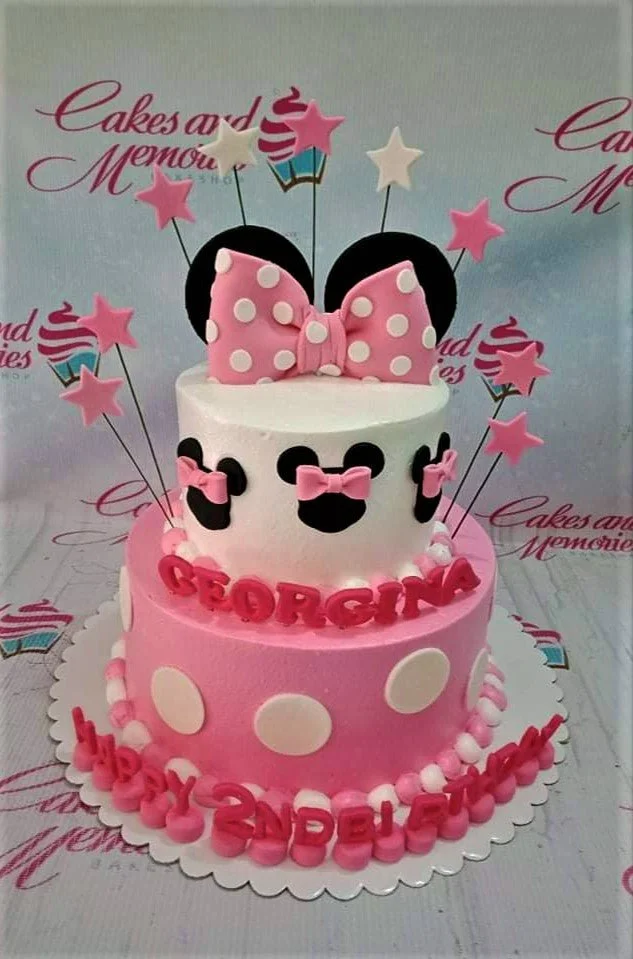 Minnie Mouse-Inspired Bow 2-Tier Birthday Cake A pink and white two-tier Minnie Mouse cake, featuring a large pink polka dot bow, black ears, and star toppers.