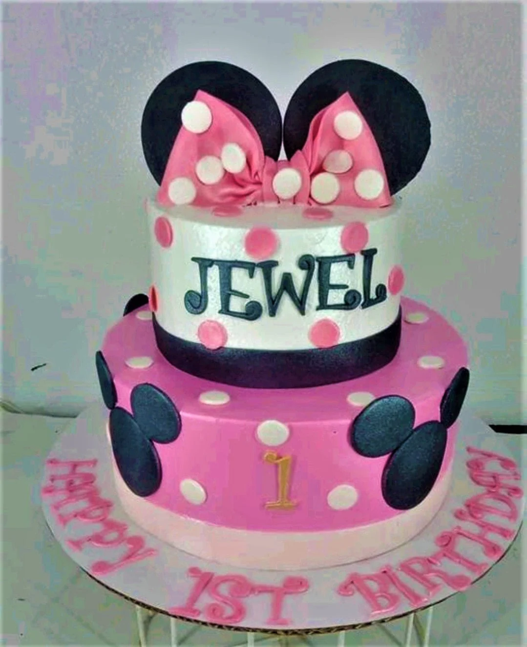 Two-tier Minnie Mouse themed birthday cake in pink, white, and black with a large pink bow and Mickey silhouettes.