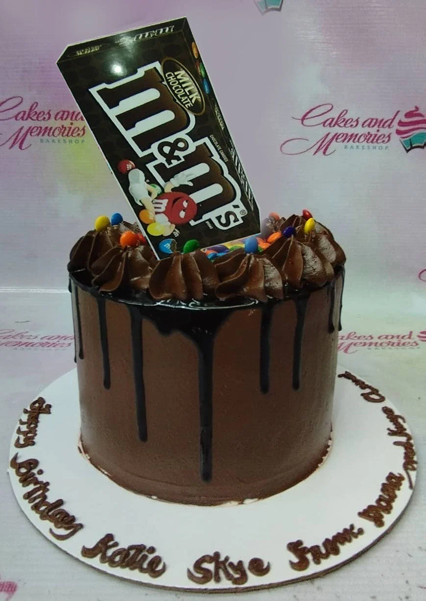 A brown 1-tier M&M's Chocolate cake, featuring an M&M's packet printout, colorful candies, and chocolate rosettes. A brown 1-tier M&M's Chocolate cake, featuring an M&M's packet printout, colorful candies, and chocolate rosettes.