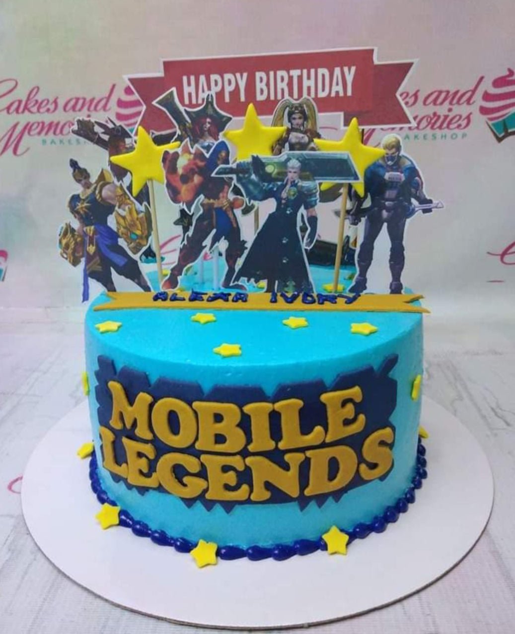 Blue Mobile Legends themed birthday cake with character printouts, yellow stars, and a Happy Birthday banner. Blue Mobile Legends themed birthday cake with character printouts, yellow stars, and a Happy Birthday banner.