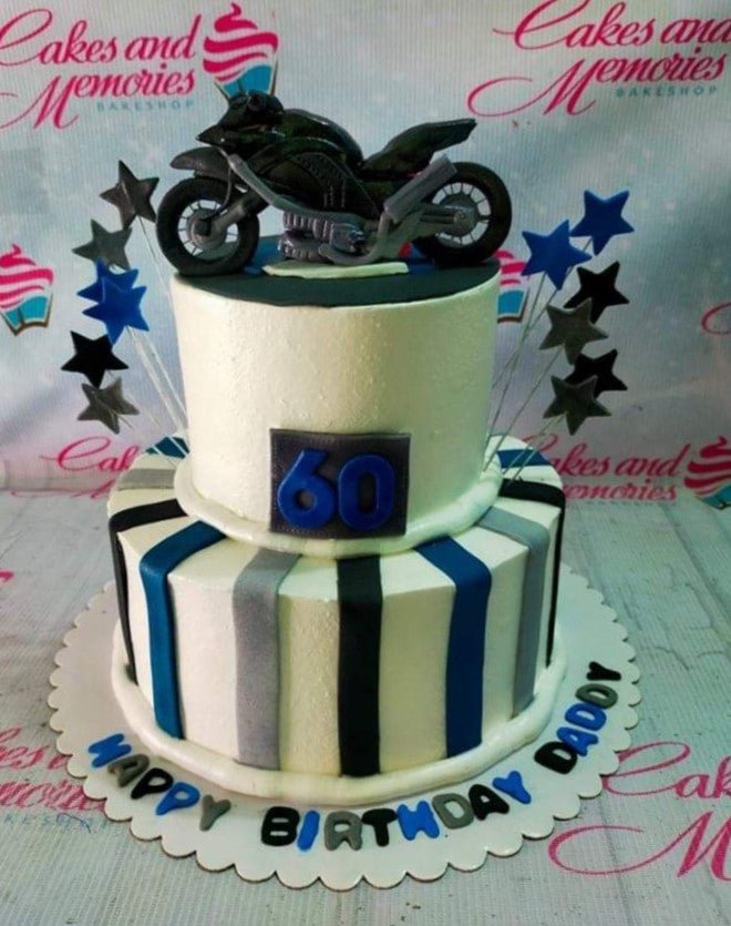 Two-tier white fondant cake with blue, gray, and black vertical stripes, topped with a detailed black motorcycle figurine and star toppers. Two-tier white fondant cake with blue, gray, and black vertical stripes, topped with a detailed black motorcycle figurine and star toppers.